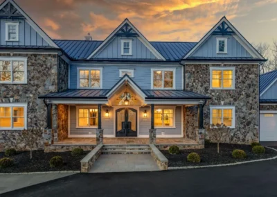 A stunning modern home with a stone facade and blue siding, featuring a grand entrance and warm lights, set against a colorful sunset