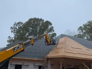 A yellow JCB telehandler extending its boom toward a partially finished residential roof covered in black underlayment