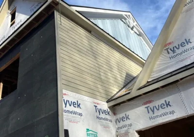 Close-up of a house under construction showing dark house wrap, yellow horizontal siding, light blue vertical siding in the gable