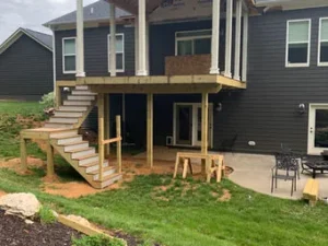Deck and stair construction on the back of a dark-sided suburban house, showing exposed wood framing and a concrete patio below