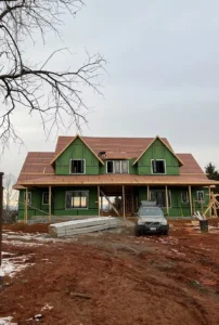 Front view of a large two-story house under construction on a muddy lot, wrapped in green house wrap
